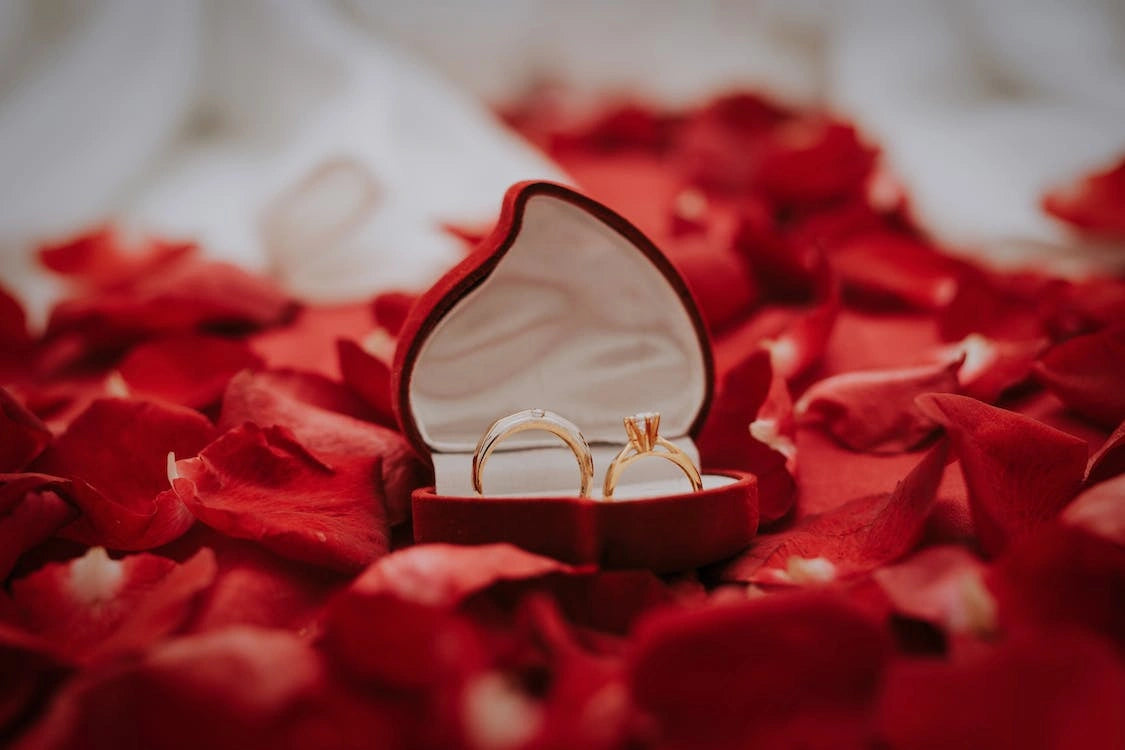 A two goldish diamond engagement rings inside a heart shaped ring box placed on  a glamorous red rose petals